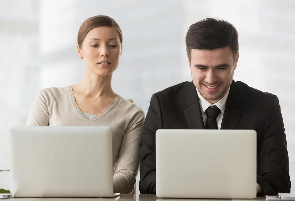 A woman looks at a man's computer to see what he is writing, exhibiting one of the common types of plagiarism