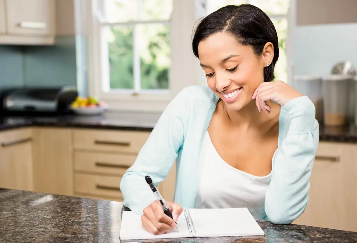 A woman sits at her kitchen counter with a notebook and pen, tracking her time for her time management plan.