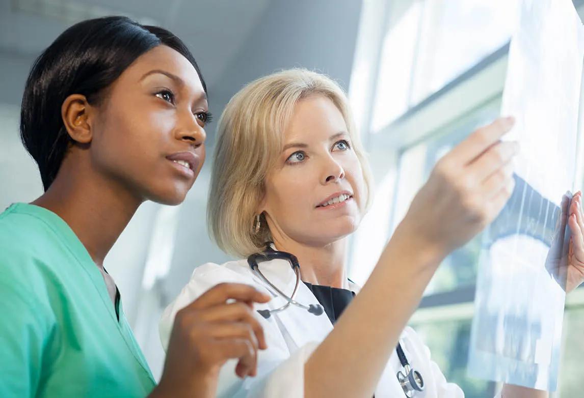 Arms of one medical administrative assistant holding a tablet next to the arms of another medical administrative assistant holding a book